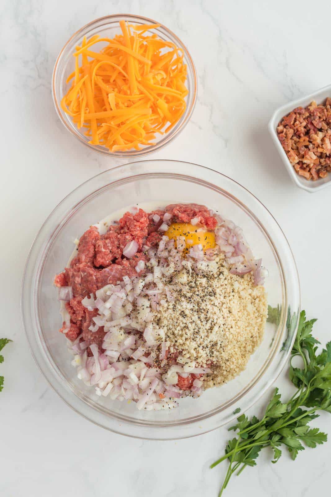 A glass bowl with ground meat, egg, breadcrumbs, onions, and spices. Nearby are bowls with shredded cheese and chopped ingredients. Parsley on the side.