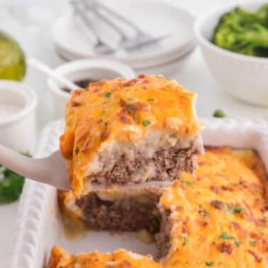 A slice of meatloaf topped with mashed potatoes and melted cheese is lifted from a baking dish. Plates, utensils, and a bowl of broccoli are in the background.