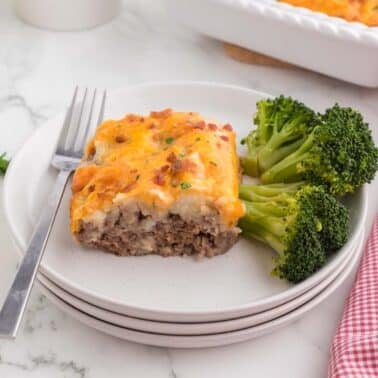 A slice of casserole with a meat base, topped with mashed potatoes and melted cheese, served with broccoli on a white plate. A fork is placed on the plate.