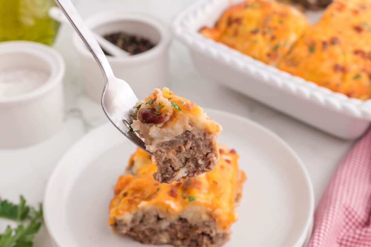 Close-up of a fork holding a bite of a casserole with visible layers of cheese and meat, over a plate and a baking dish with more casserole in the background.