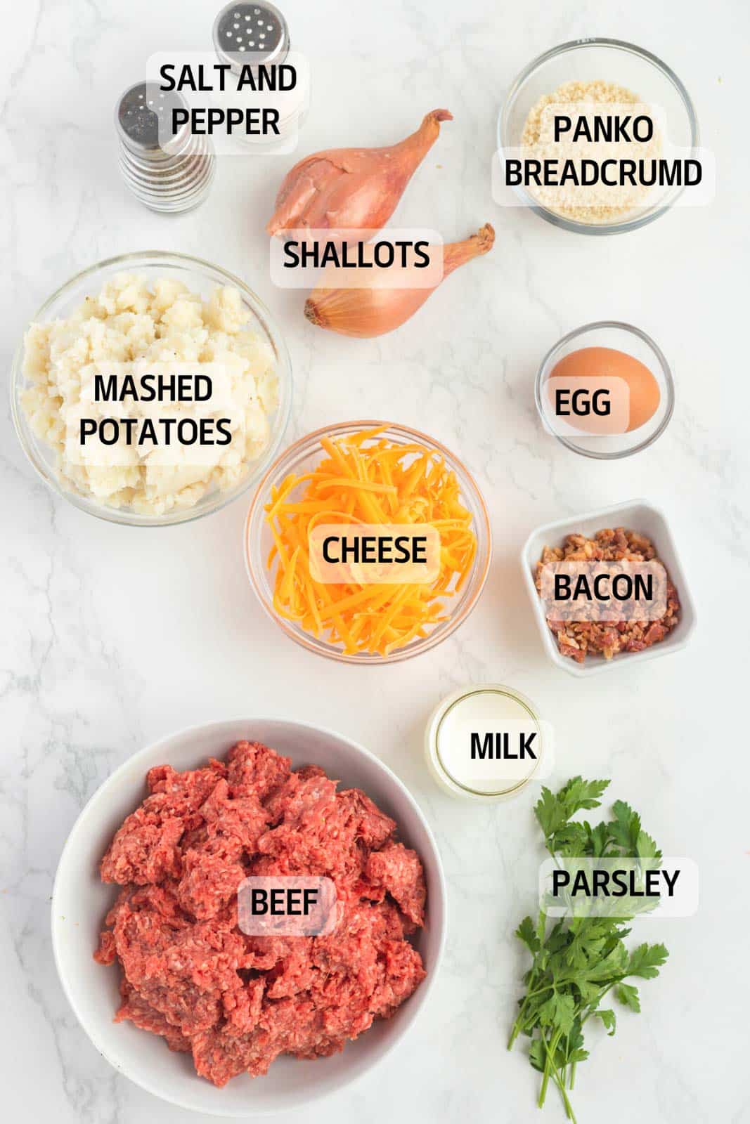 Ingredients for a dish arranged on a countertop include ground beef, mashed potatoes, shallots, cheese, panko breadcrumbs, egg, bacon, milk, parsley, with salt and pepper.