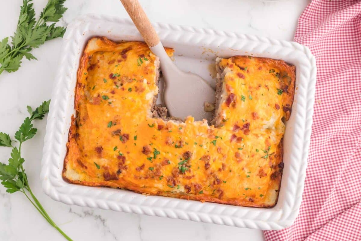 A casserole dish with a portion removed, revealing layers of meat and cheese, topped with a golden crust. A serving spatula rests in the dish. Parsley and a red checkered cloth are nearby.