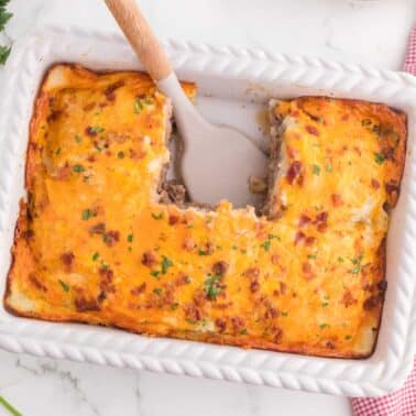 A casserole dish with a portion removed, revealing layers of meat and cheese, topped with a golden crust. A serving spatula rests in the dish. Parsley and a red checkered cloth are nearby.