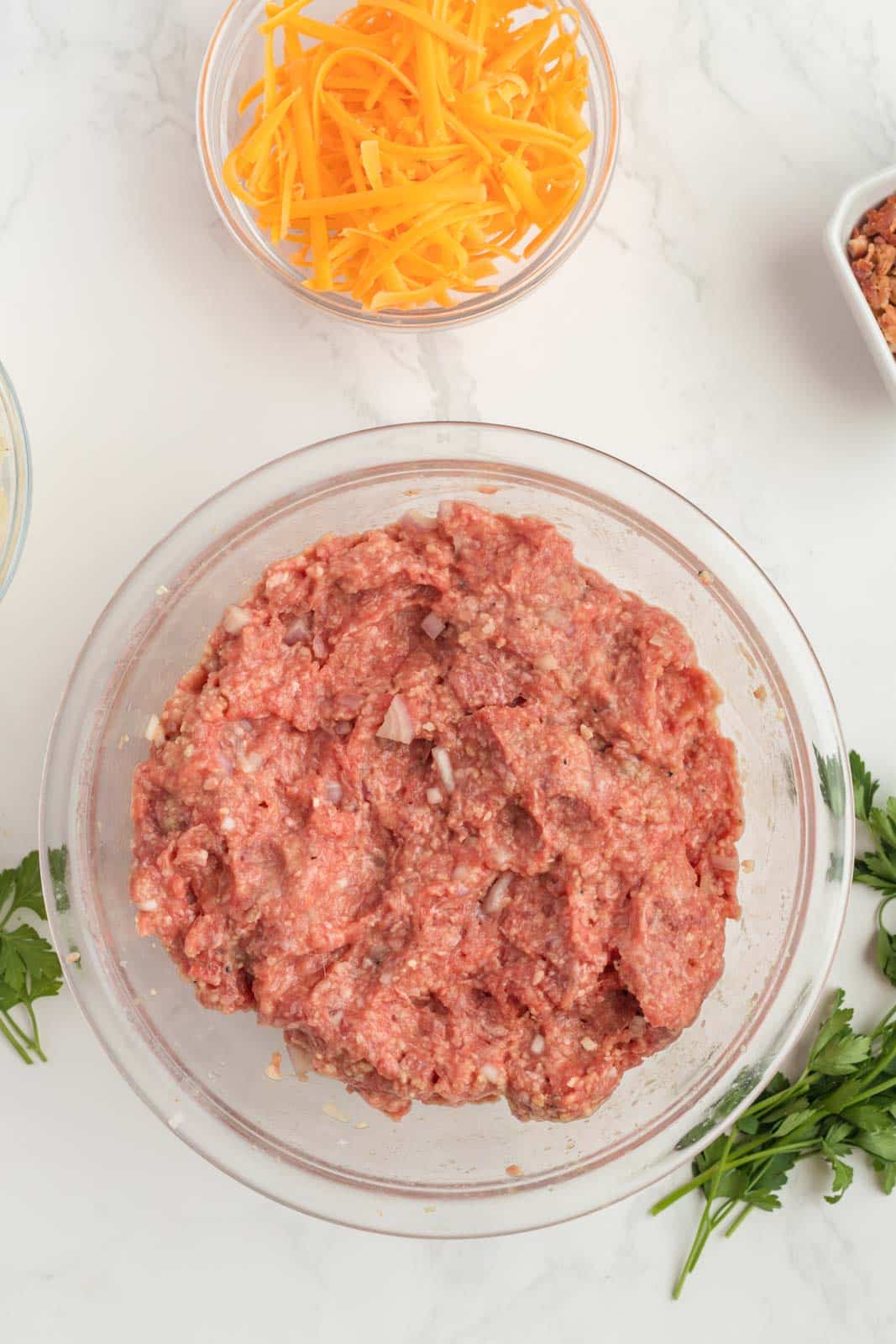 Ground meat mixture in a glass bowl, surrounded by grated cheese, chopped parsley, and a small dish of bacon pieces on a white surface.