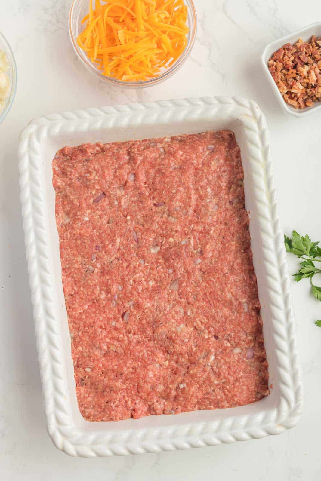 Ground meat evenly spread in a rectangular baking dish, surrounded by small bowls of grated cheese, bacon bits, and parsley on a marble surface.