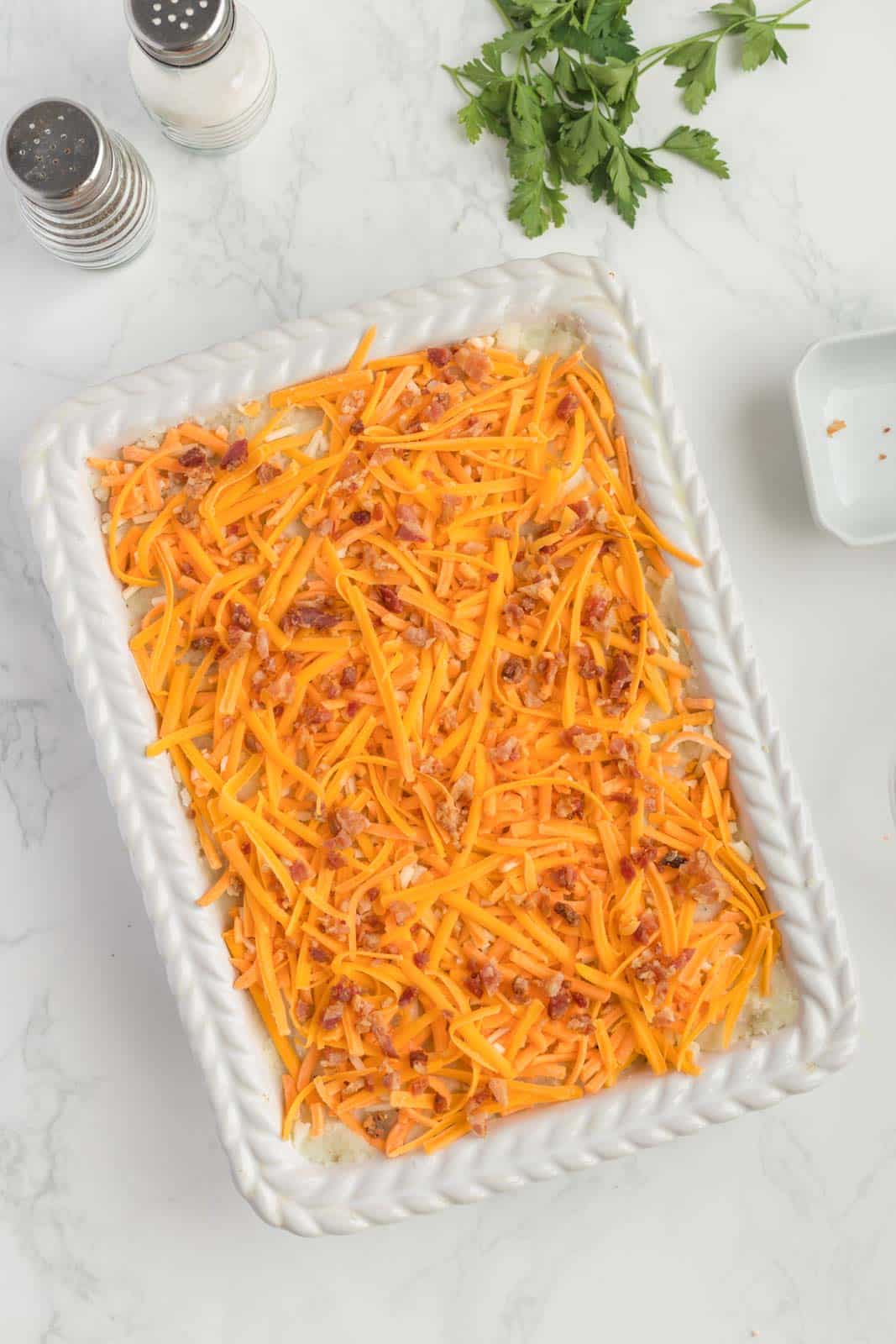 Rectangular dish with shredded cheddar cheese on top of a layered casserole. Parsley, a salt shaker, and a pepper shaker are nearby on a white marble surface.