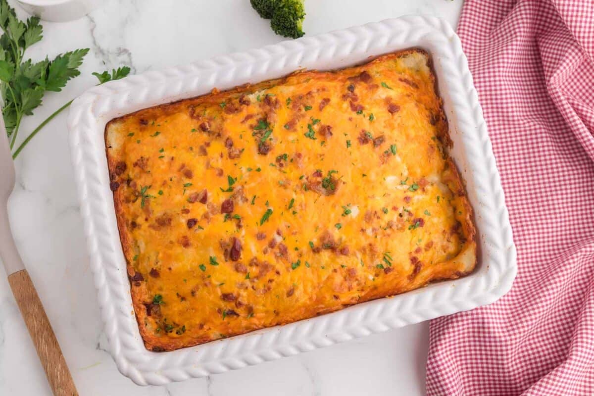 A baked casserole with a golden cheese topping in a white dish on a marble surface, accompanied by parsley, broccoli, and a red checkered cloth.