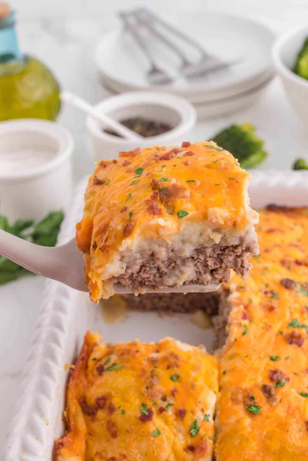 A spatula lifts a slice of meat and mashed potato casserole topped with melted cheese and herbs from a white baking dish.