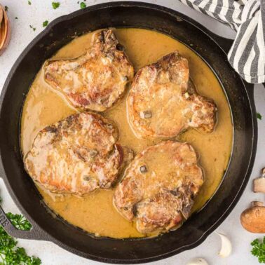 Pork chops in a creamy sauce are cooking in a black skillet, surrounded by sliced mushrooms and fresh parsley on a white surface.