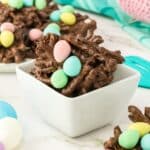 A white bowl filled with chocolate-covered pretzel clusters, topped with pastel-colored egg-shaped candies, on a marble surface.
