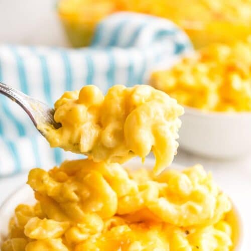 A fork holds a bite of creamy macaroni and cheese above a bowl, with another full bowl in the background next to a striped cloth.