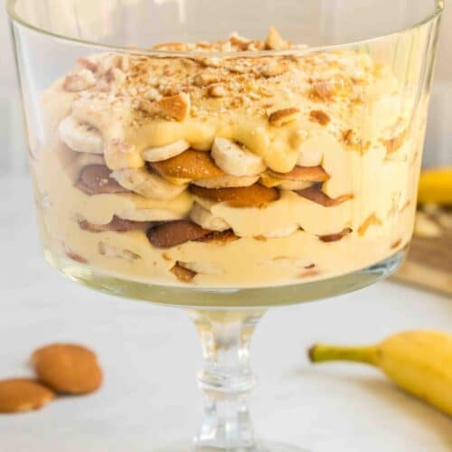 A glass trifle bowl filled with layers of banana pudding, vanilla wafers, and sliced bananas, topped with crumbled wafers.