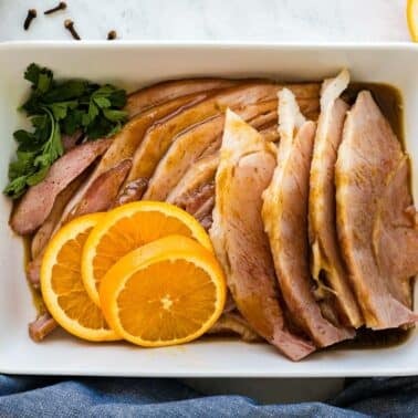 Sliced bourbon glazed ham arranged in a rectangular dish, garnished with parsley and orange slices, with a blue cloth napkin beside it.