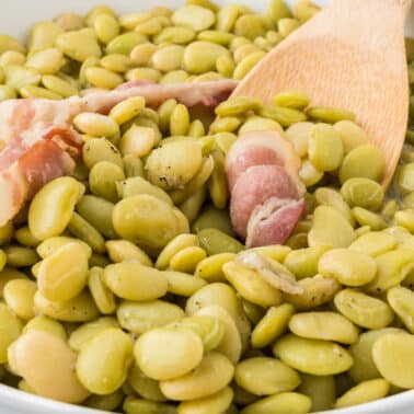 A bowl of cooked lima beans with pieces of bacon and a wooden spoon.