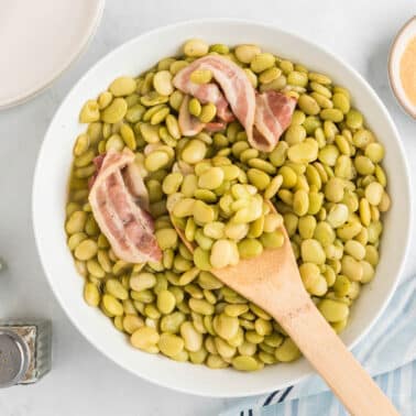 Bowl of lima beans with bacon slices, beside a dish of cornmeal and salt and pepper shakers, with a wooden spoon and striped cloth on a white surface.