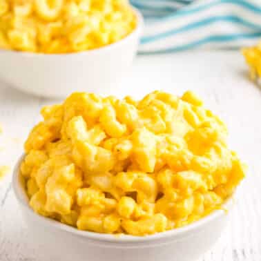 A bowl of creamy macaroni and cheese with a bright yellow color sits on a white surface. A striped blue and white cloth is in the background, and a fork rests nearby.