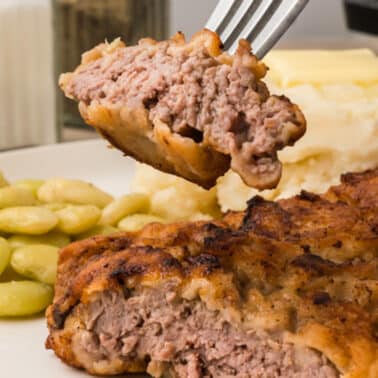 Close-up of a fork holding a piece of breaded cubed steak. Plate includes mashed potatoes with butter and lima beans in the background.