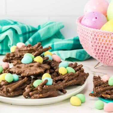 Chocolate clusters topped with colorful candy eggs are on a white plate. In the background, a pink basket holds pastel Easter eggs. A turquoise cloth is draped nearby.