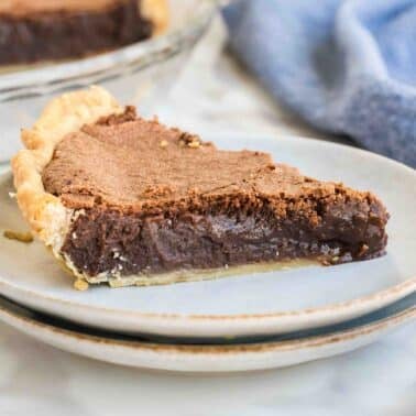 A slice of fudge pie on a stack of two plates, with a golden flaky crust and rich, creamy filling, set on a marble surface. A blue cloth is in the background.