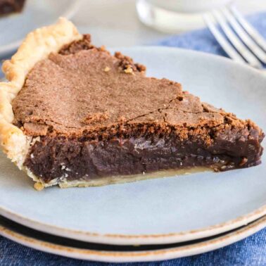 Slice of chocolate fudge pie on a light blue plate, placed on a blue cloth, with a fork in the background.