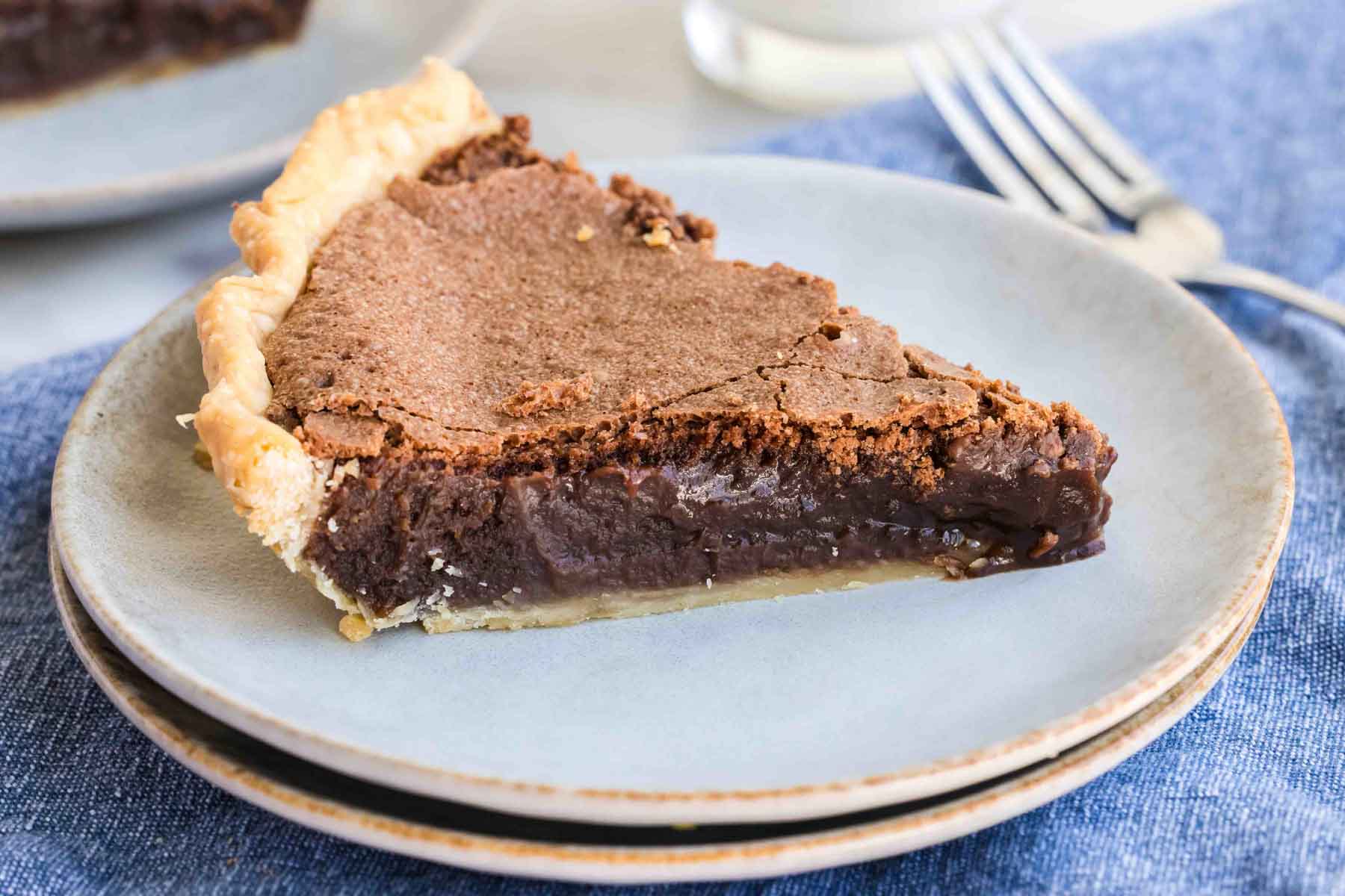 Slice of chocolate fudge pie on a light blue plate, placed on a blue cloth, with a fork in the background.
