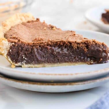 A slice of fudge pie sits on a white plate, revealing a rich, creamy filling encased in a flaky crust. Two other pieces are visible in the background.
