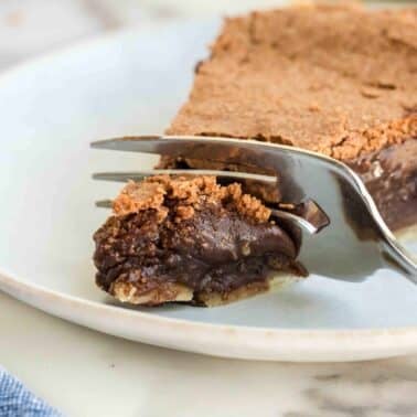 A fork cutting into a slice of fudge pie with a flaky crust on a white plate.