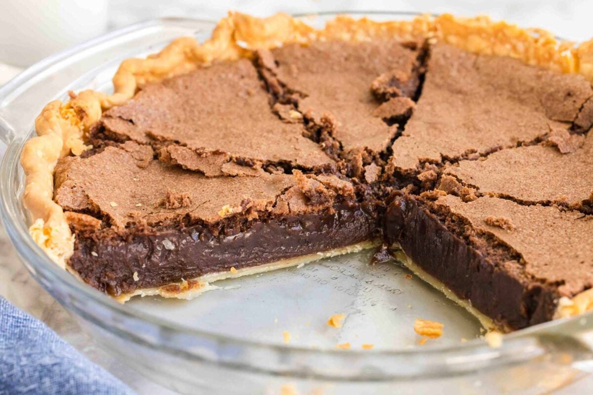 A chocolate pie in a clear glass pie dish with a slice missing, revealing a dark, rich filling and a flaky crust. Cracks are visible on the pie's surface.