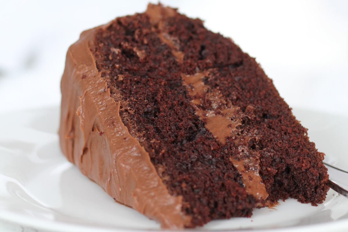 A slice of rich chocolate cake with chocolate frosting on a white plate.