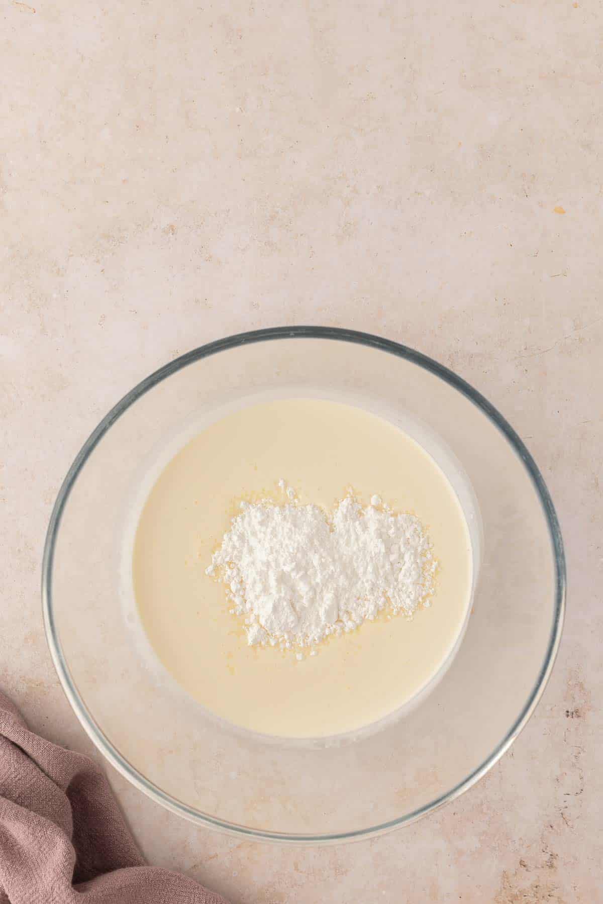 Mixing bowl with liquid batter and a heap of flour on a light countertop.