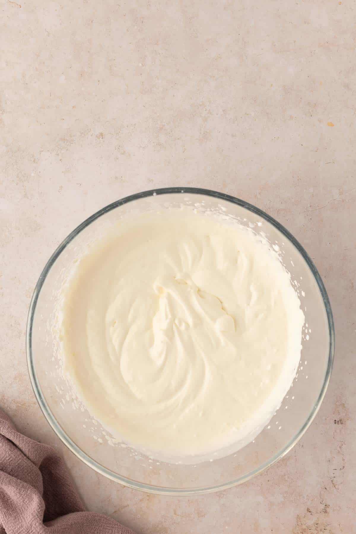 A glass bowl filled with smooth, whipped cream sits on a light marble surface. A corner of a mauve cloth is visible in the bottom left of the image.