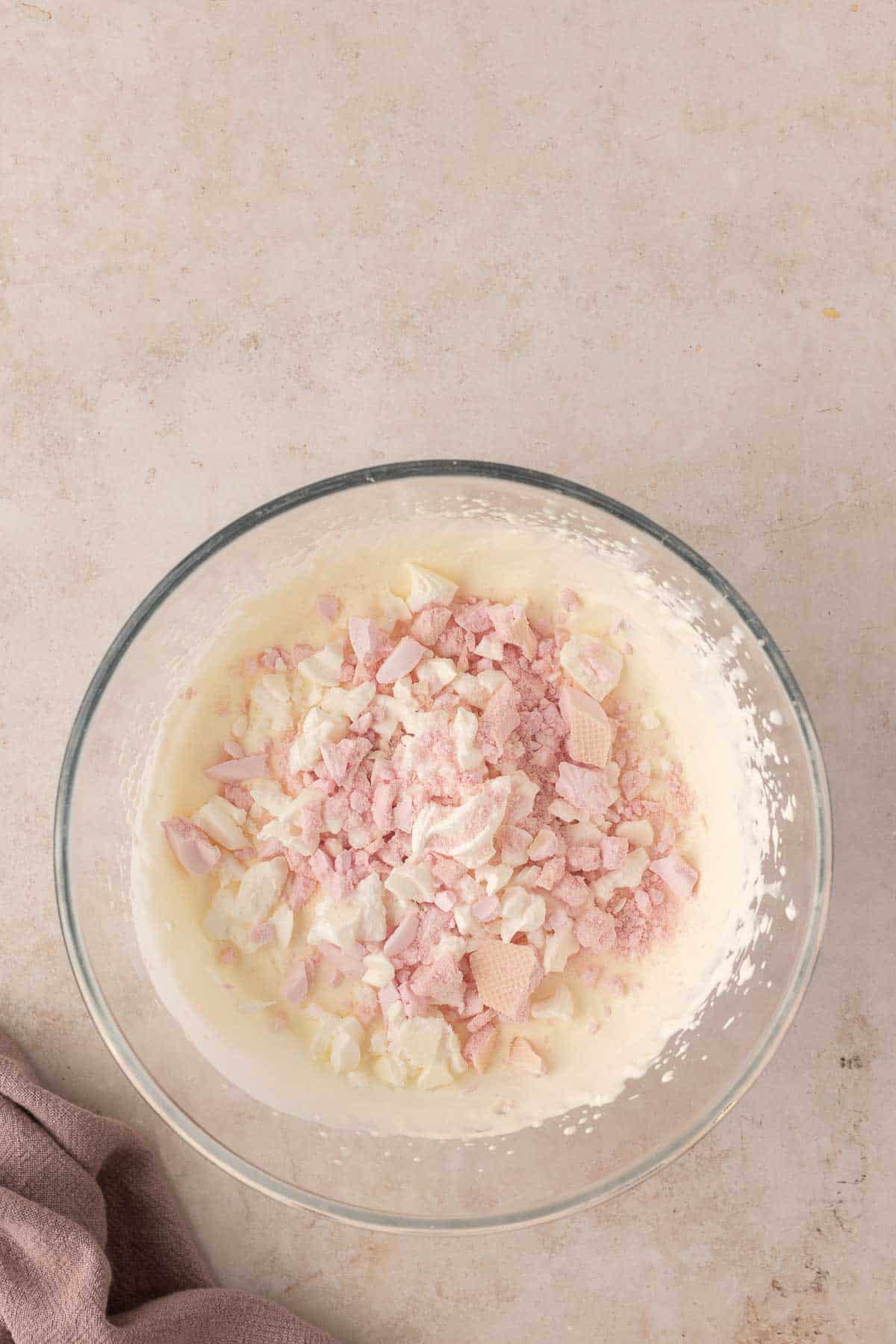 A glass bowl with a cream mixture topped with pink and white crumb-like pieces on a beige surface. A cloth is partially visible in the corner.