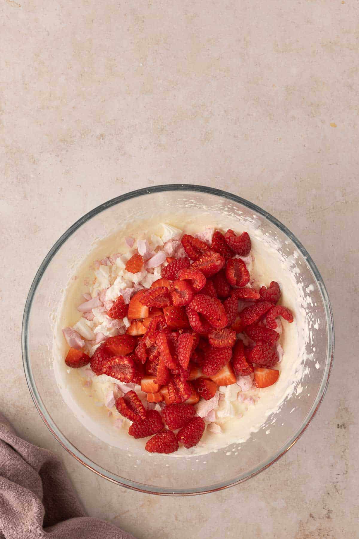 Glass bowl with creamy mixture topped with sliced strawberries, diced peaches, and small marshmallows on a light-colored surface, next to a crumpled cloth.