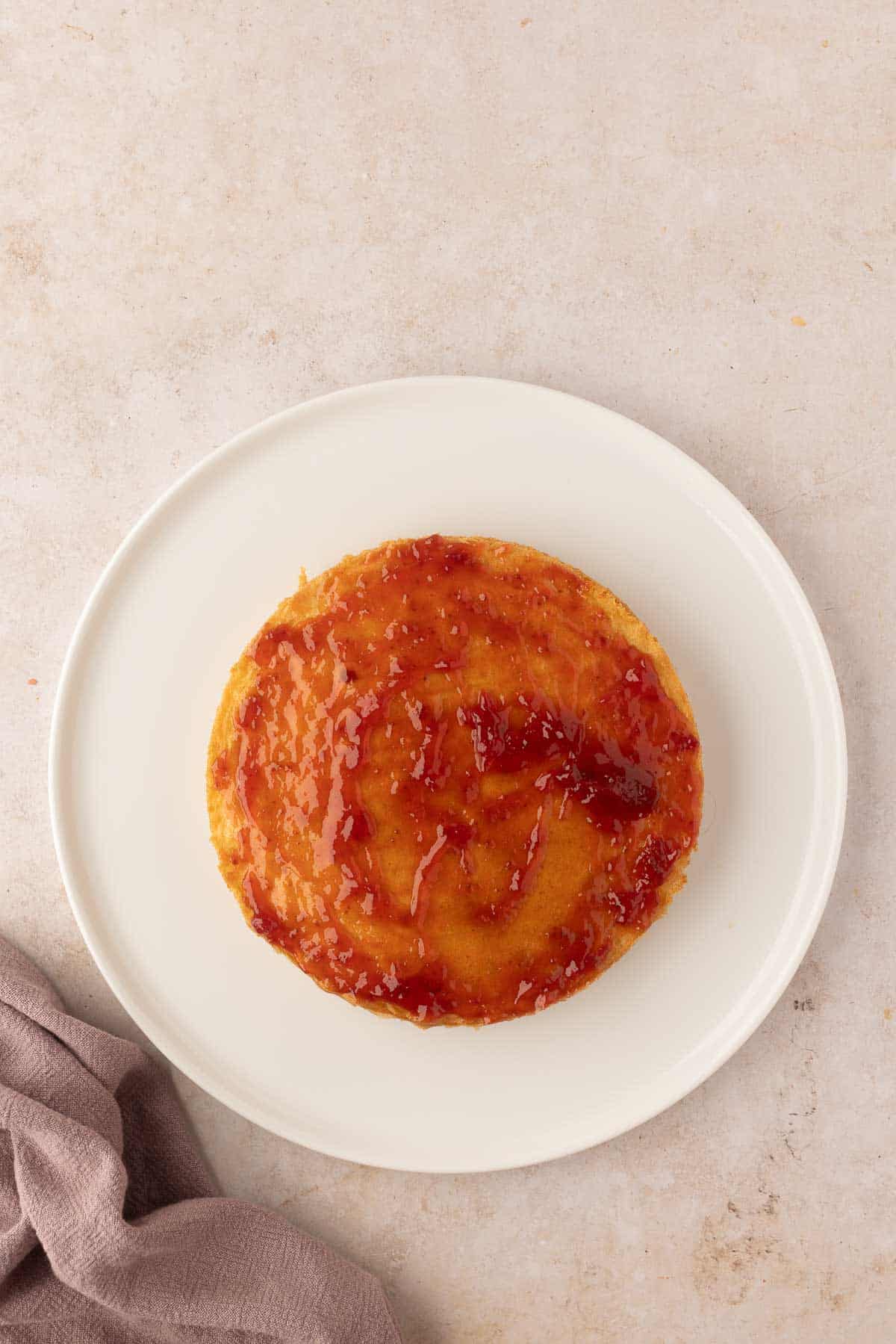 A round caramel flan on a white plate, set on a light beige surface with a folded gray cloth nearby.