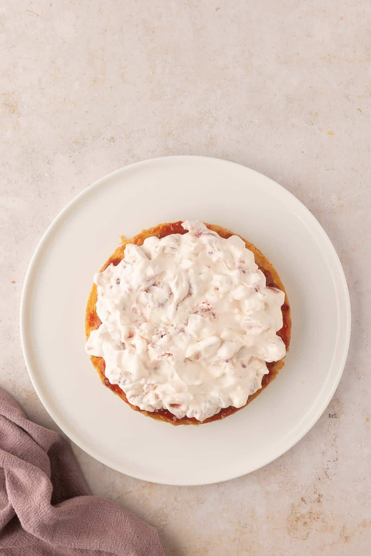 A round cake topped with cream and jam on a white plate, placed on a light beige surface with a folded napkin beside it.