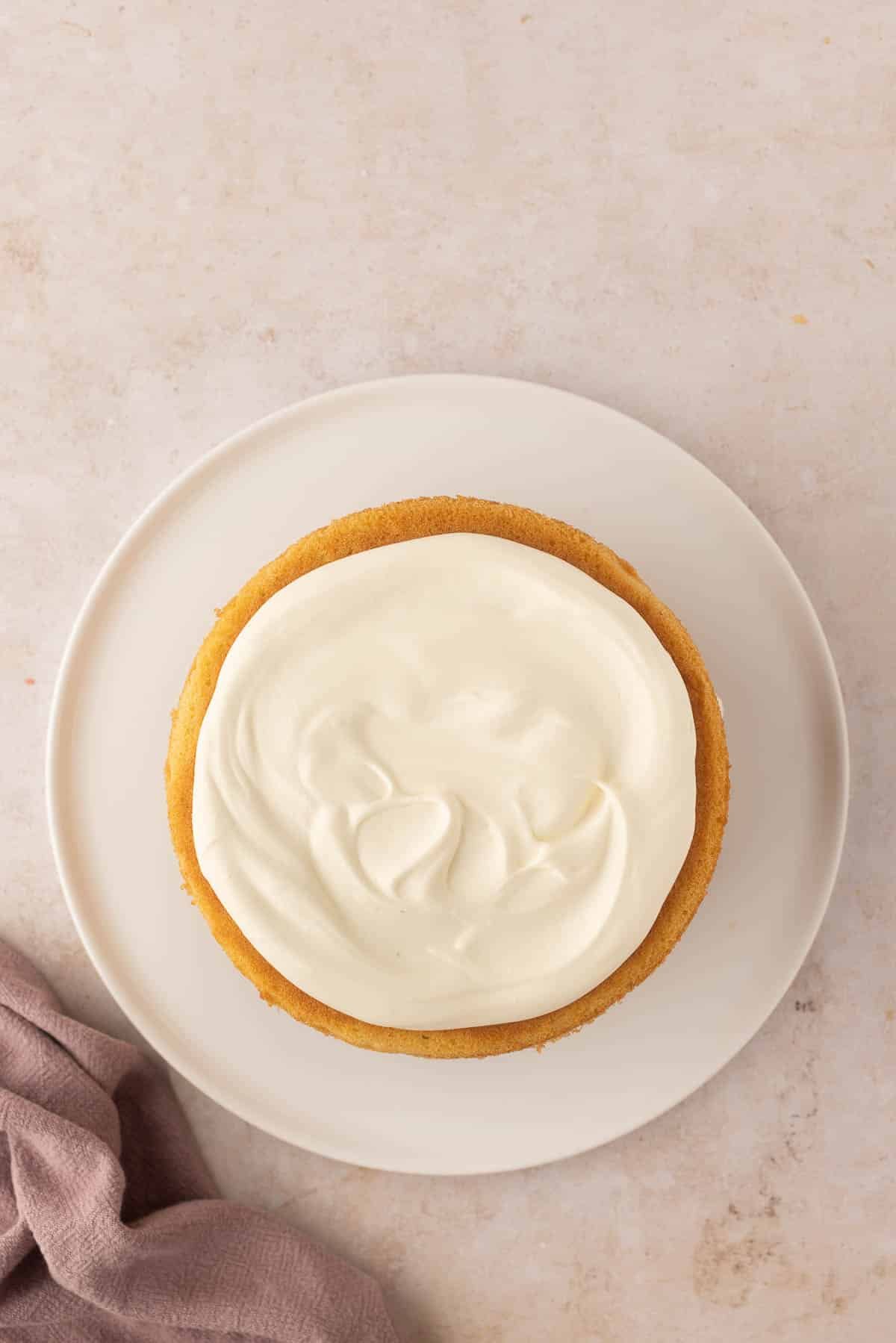 A round cake with smooth cream frosting on a white plate. A folded light purple cloth is placed next to the plate on a beige surface.