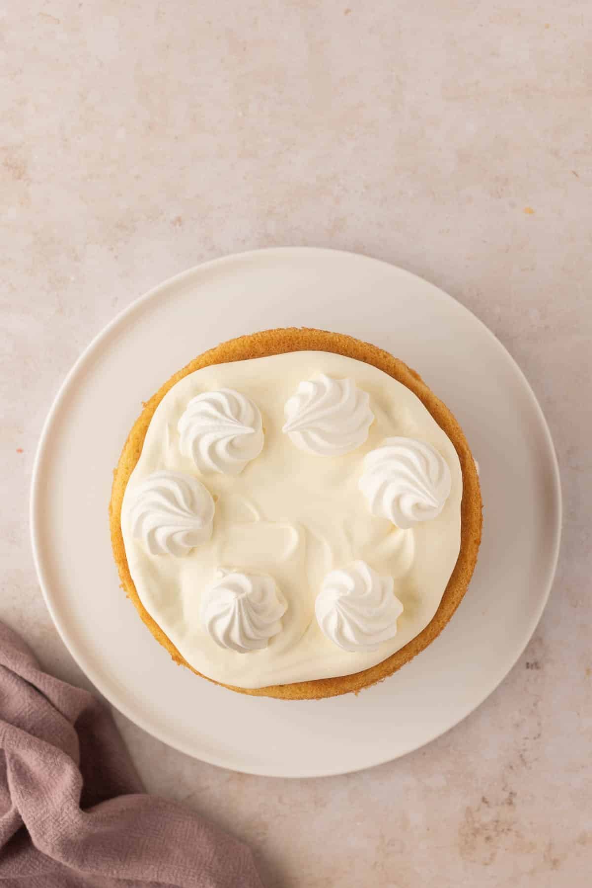 A round cake with white frosting and six dollops of cream on top, placed on a white plate on a light-colored surface. A folded cloth is partially visible on the side.