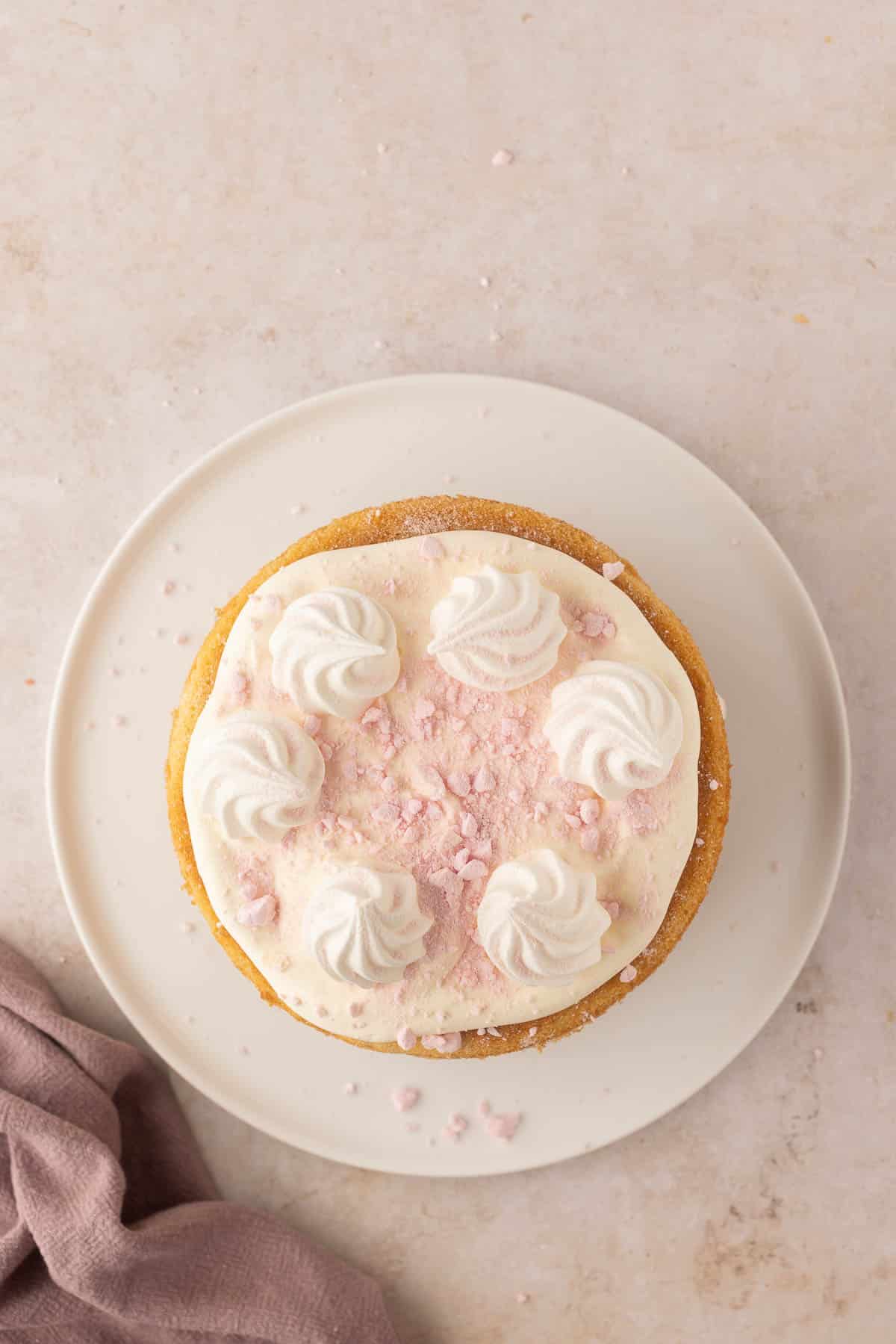 A round cake with white frosting swirls and pink crumbles on top is placed on a white plate, resting on a light-colored surface with a pink cloth nearby.