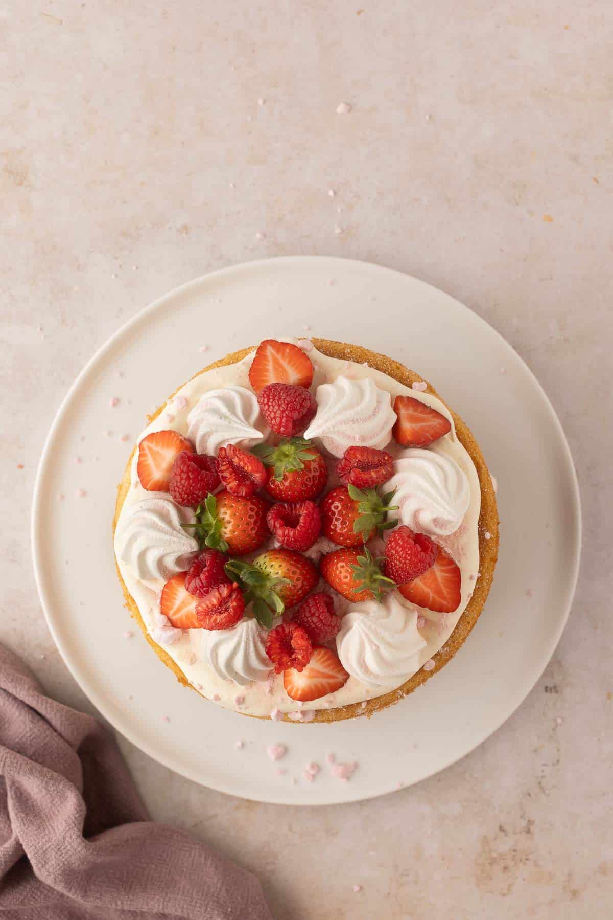 A round cake topped with whipped cream, strawberries, and raspberries on a white plate, set on a light-colored surface with a cloth nearby.