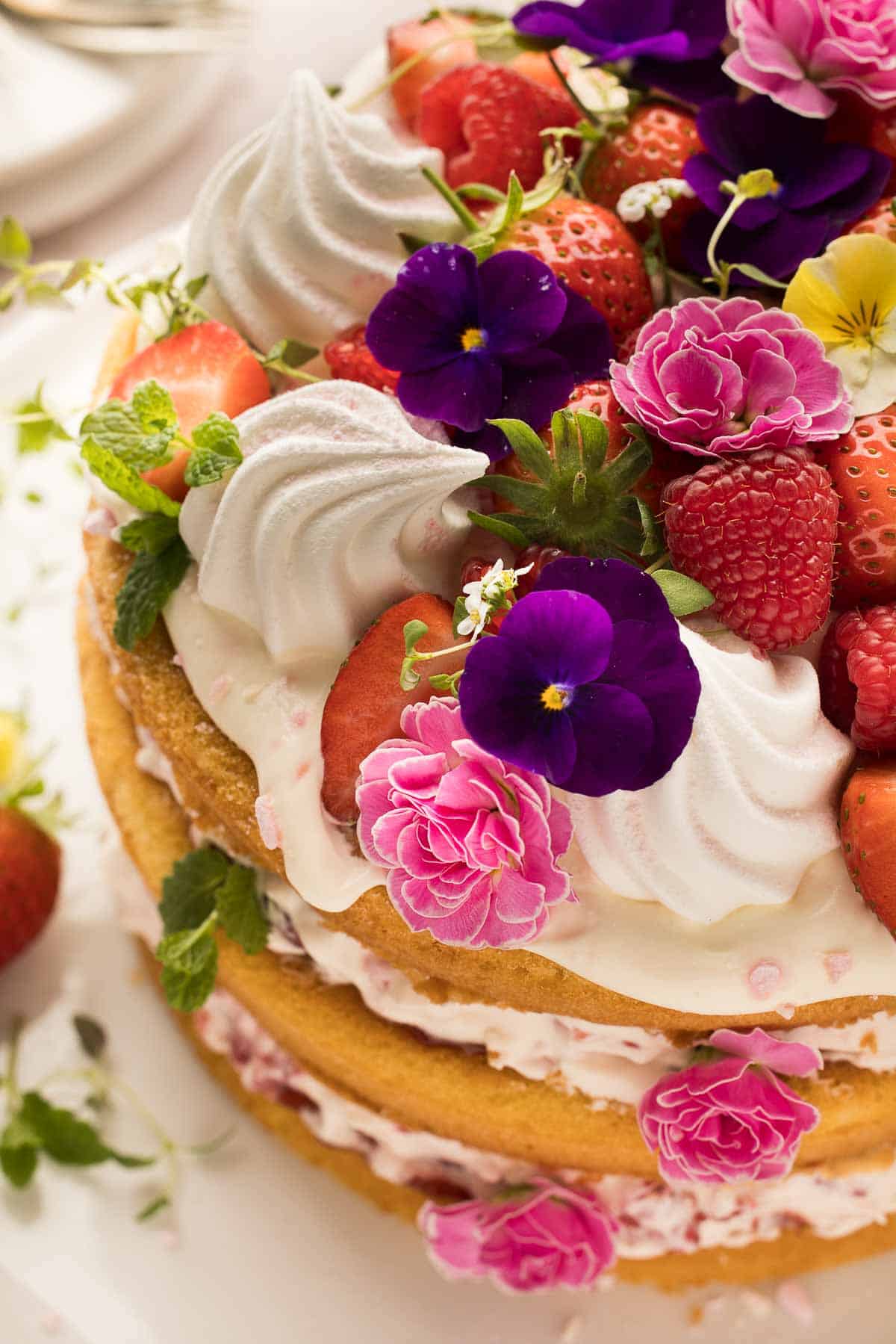 A layered Eton mess cake topped with whipped cream, strawberries, raspberries, and edible flowers, including pansies and carnations. Decorated with fresh herbs.