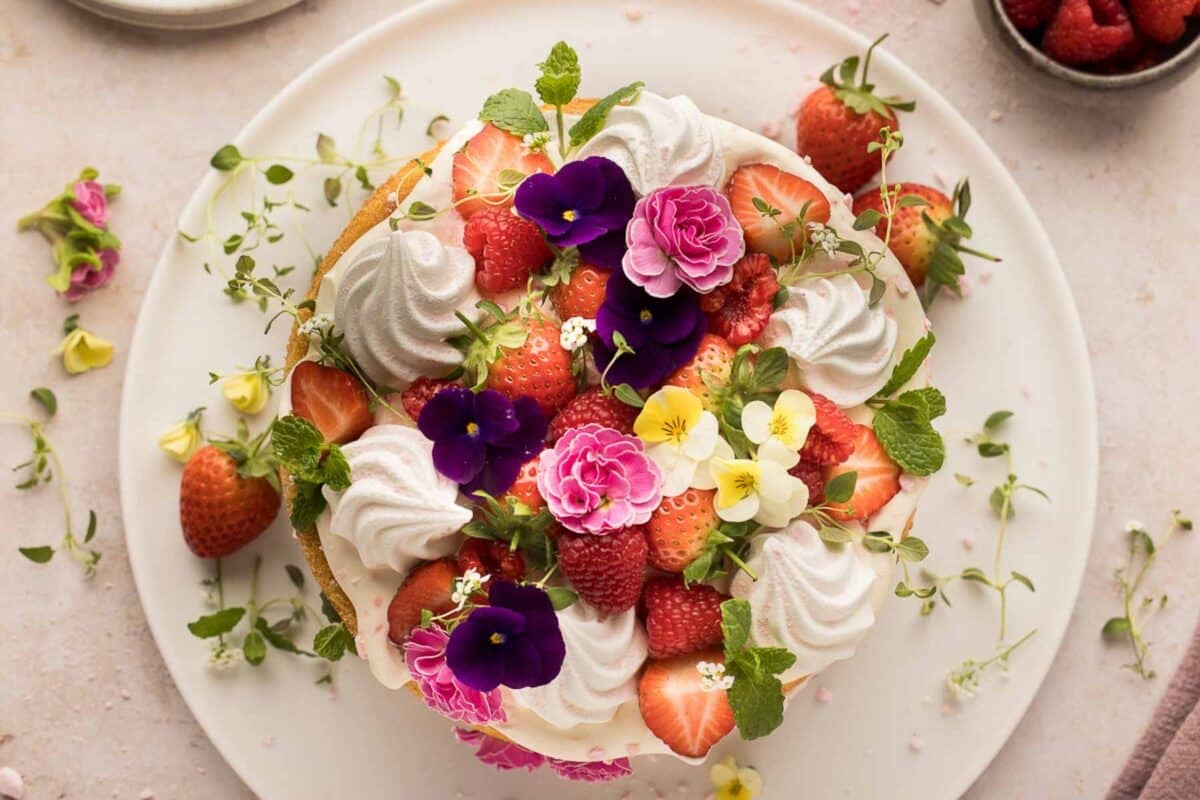 A naked cake with flowers topped with whipped cream, strawberries, raspberries, and assorted edible flowers on a white plate.