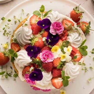 A naked cake with flowers topped with whipped cream, strawberries, raspberries, and assorted edible flowers on a white plate.