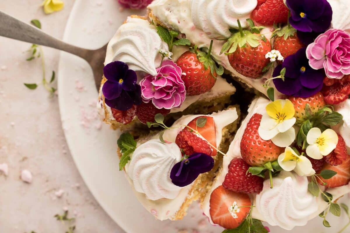 A slice of naked cake topped with strawberries, whipped cream, and colorful flowers is being lifted from the plate.