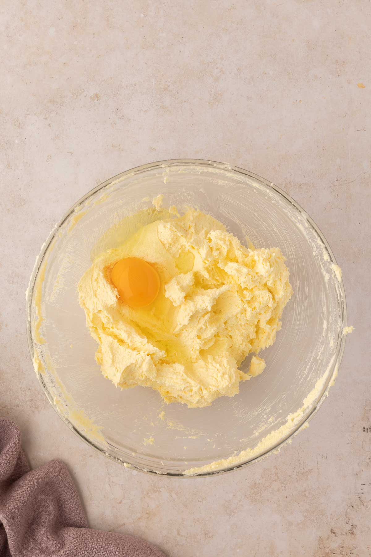 A clear glass bowl with yellow cake batter and a raw egg on a light marble surface. A brown cloth is partially visible at the lower left corner.