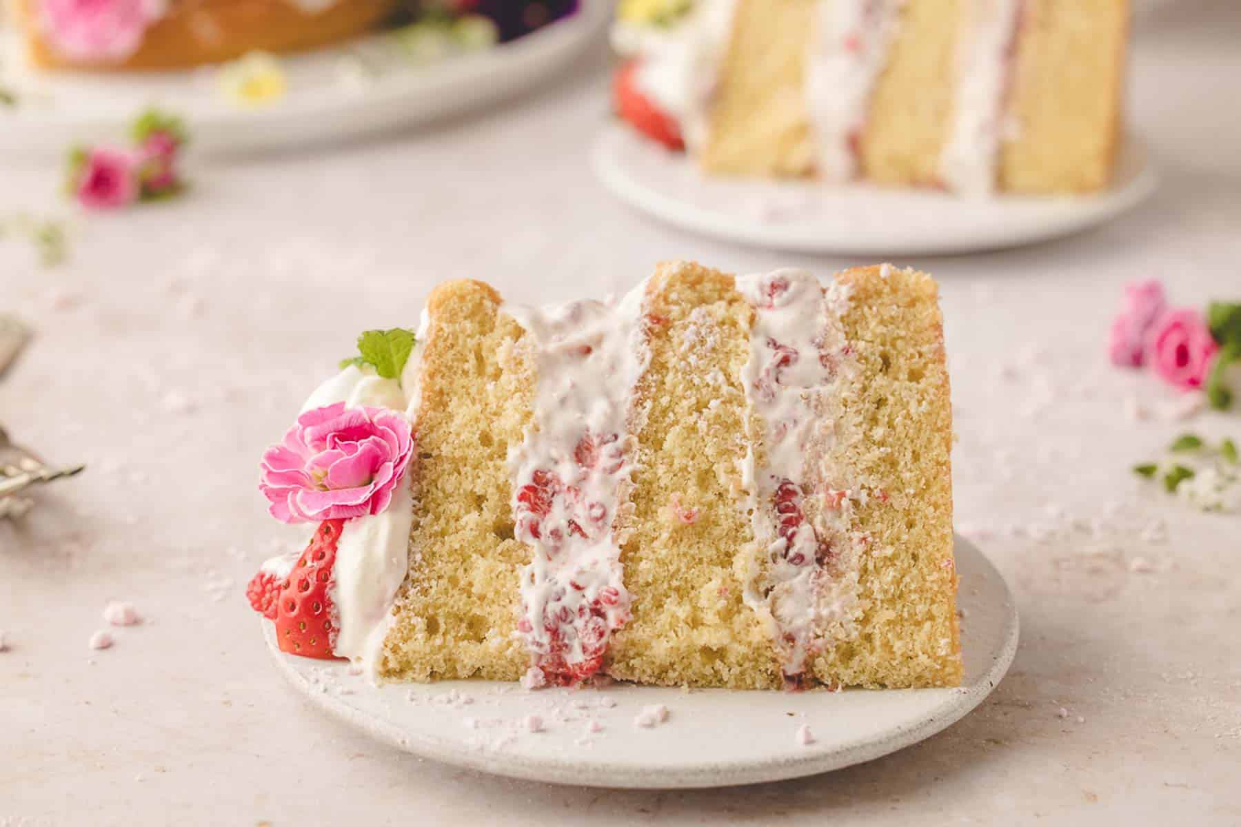 Slice of Eton mess cake with cream and strawberries, decorated with a pink flower, on a white plate.