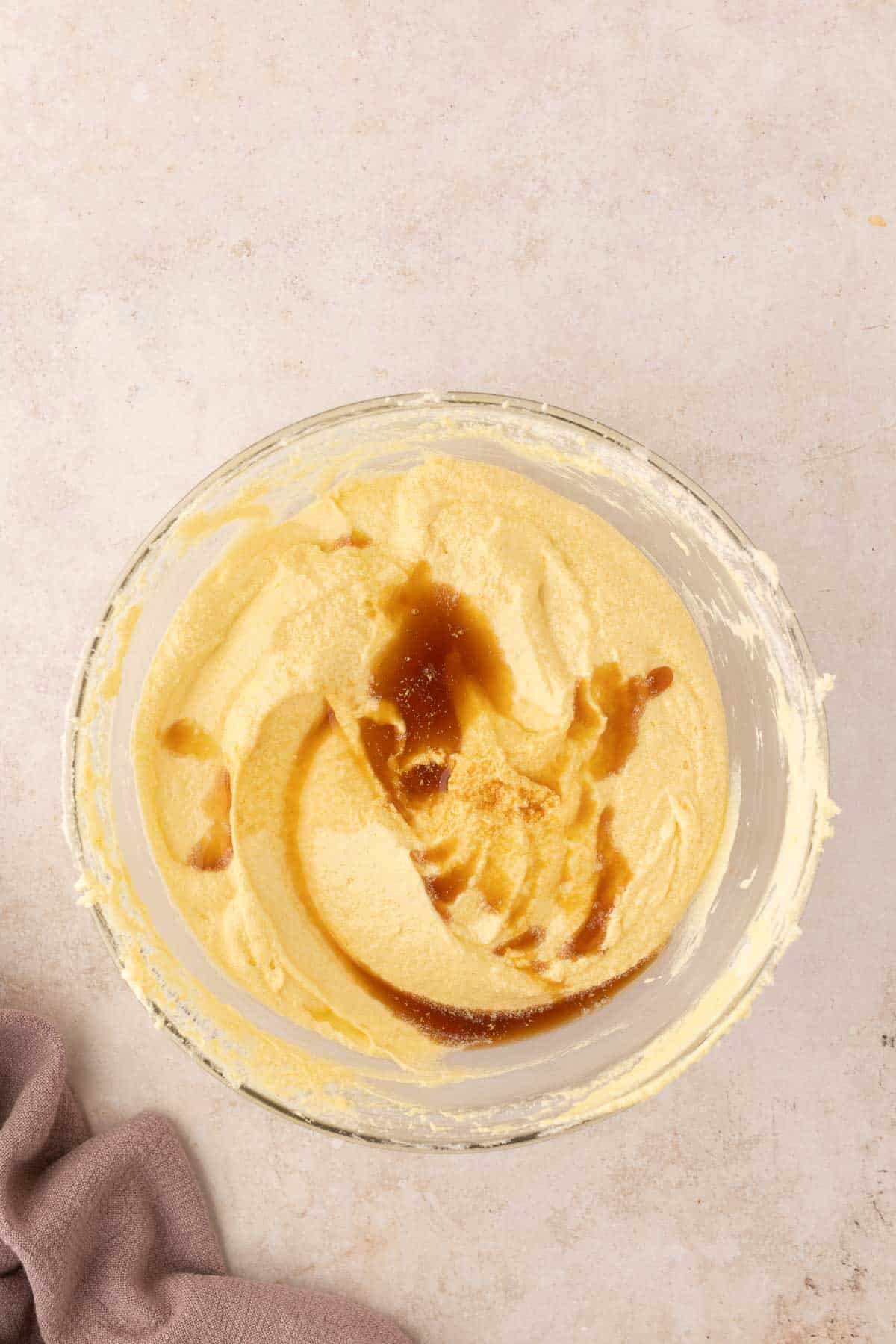 A bowl of yellow cake batter with a swirl of vanilla extract on a light surface. A purple cloth is partially visible on the side.