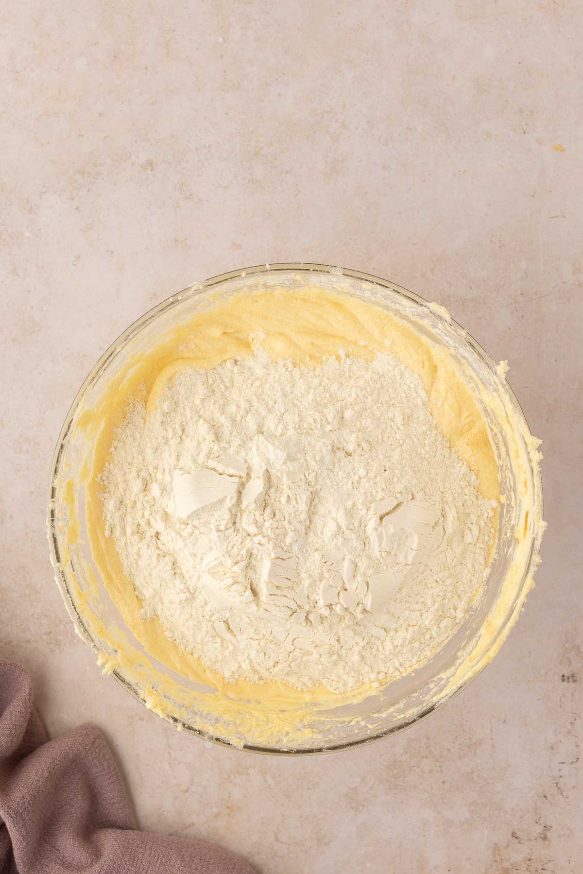 A glass bowl with yellow cake batter and flour on top, placed on a light countertop. A brown cloth is partially visible on the left.