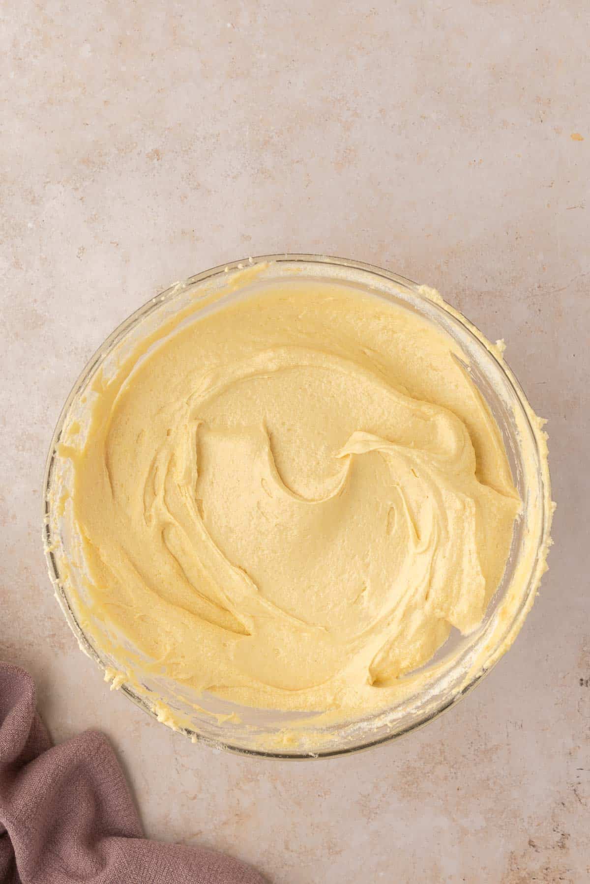 A glass bowl filled with smooth, creamy yellow cake batter on a beige surface, with a folded brown cloth nearby.