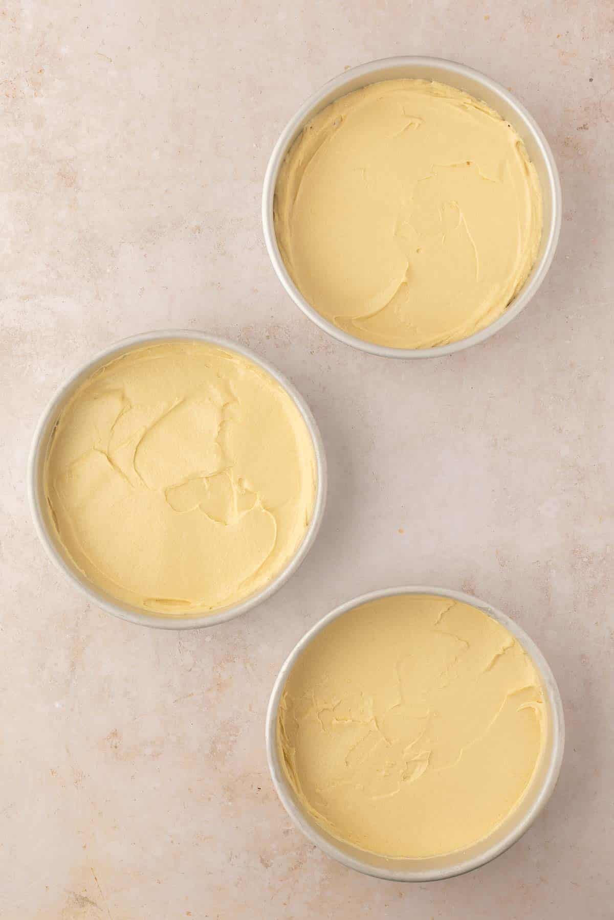 Three round baking pans filled with smooth, light yellow cake batter placed on a beige textured surface.