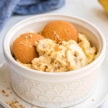 A white bowl of Southern banana pudding topped with two vanilla wafers and banana slices, with a few crumbs scattered around, placed on a white saucer. A banana and a cloth are in the background.